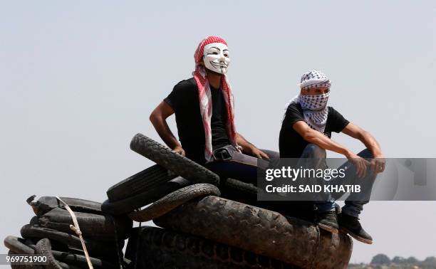 Palestinian protester wearing a Guy Fawkes mask sits on tires next to another covering his face with a keffiyeh during a demonstration near the...