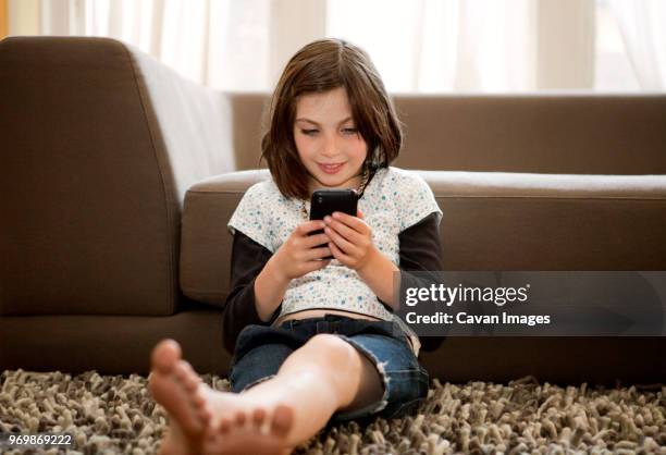 happy girl using mobile phone while sitting against sofa - happy mobile stockfoto's en -beelden