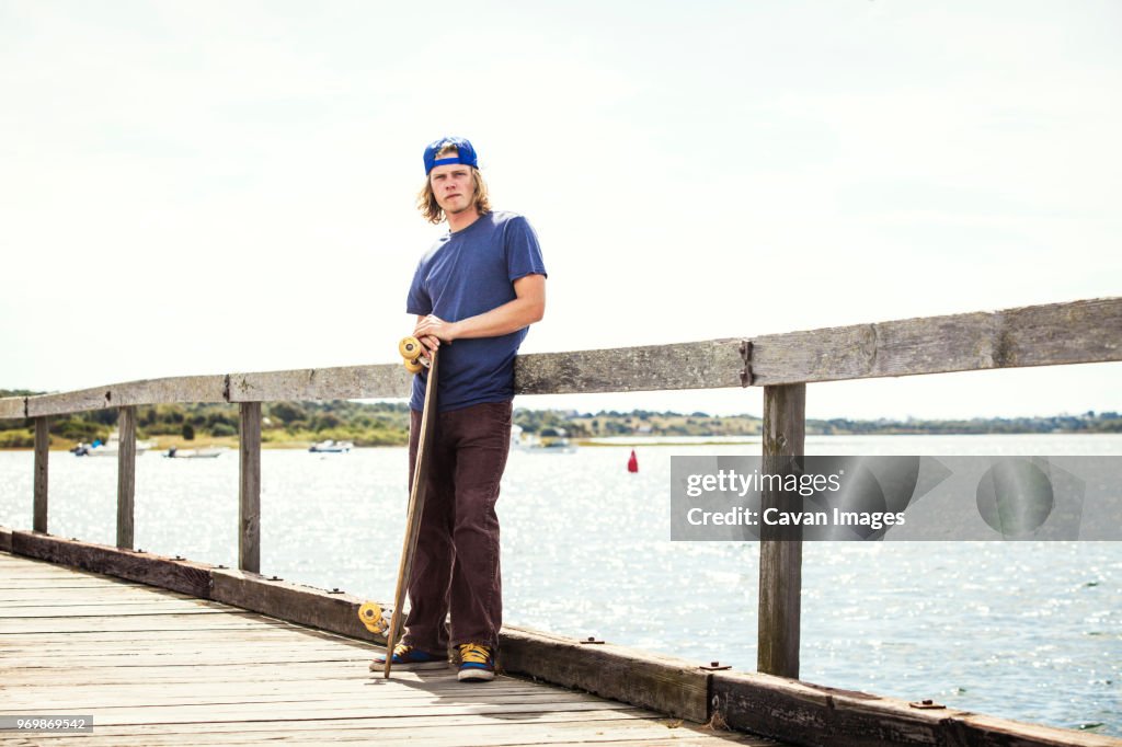 Portrait of man with longboard standing on pier against clear sky
