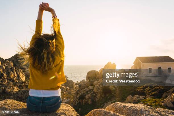 italy, sardinia, woman on a hiking trip sitting on rock at the coast raising her arms - rock terrain stockfoto's en -beelden
