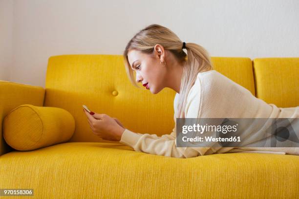 blonde woman lying on sofa, using smartphone at home - acostado fotografías e imágenes de stock