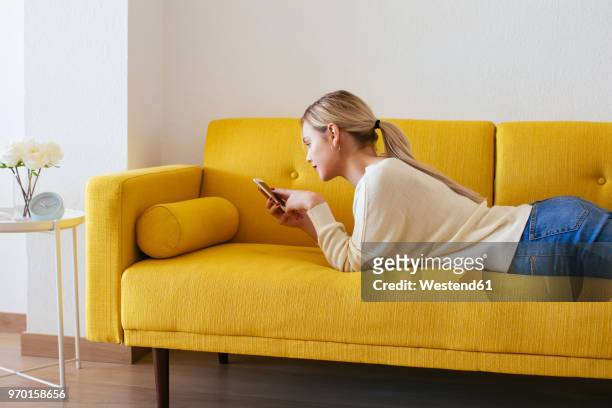blonde woman lying on sofa, using smartphone at home - junge frau uhr stock-fotos und bilder