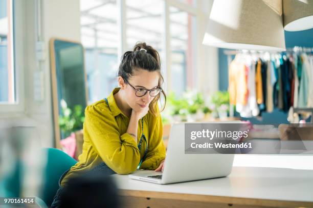 fashion designer sitting at desk in her studio looking at laptop - ontwerp beroep stockfoto's en -beelden