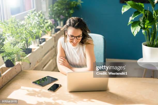 smiling freelancer sitting at desk in loft looking at laptop - sitting and using smartphone studio stockfoto's en -beelden