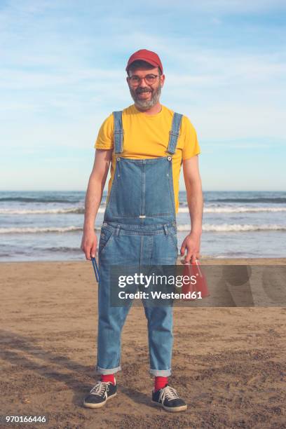 portrait of smiling man in dungarees standing on the beach holding bottles of soft drinks - dungarees stock-fotos und bilder