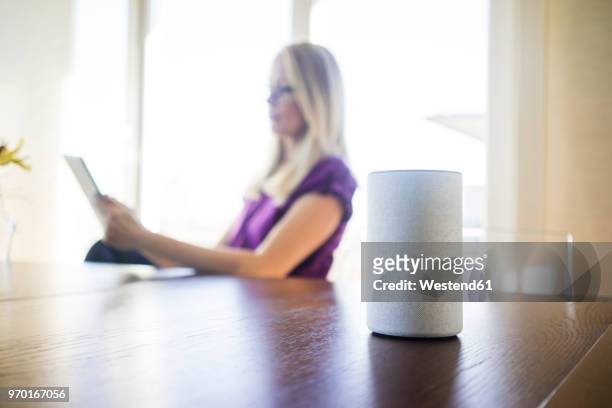 smart home loudspeaker on table with woman using tablet in the background - bluetooth speaker stock-fotos und bilder