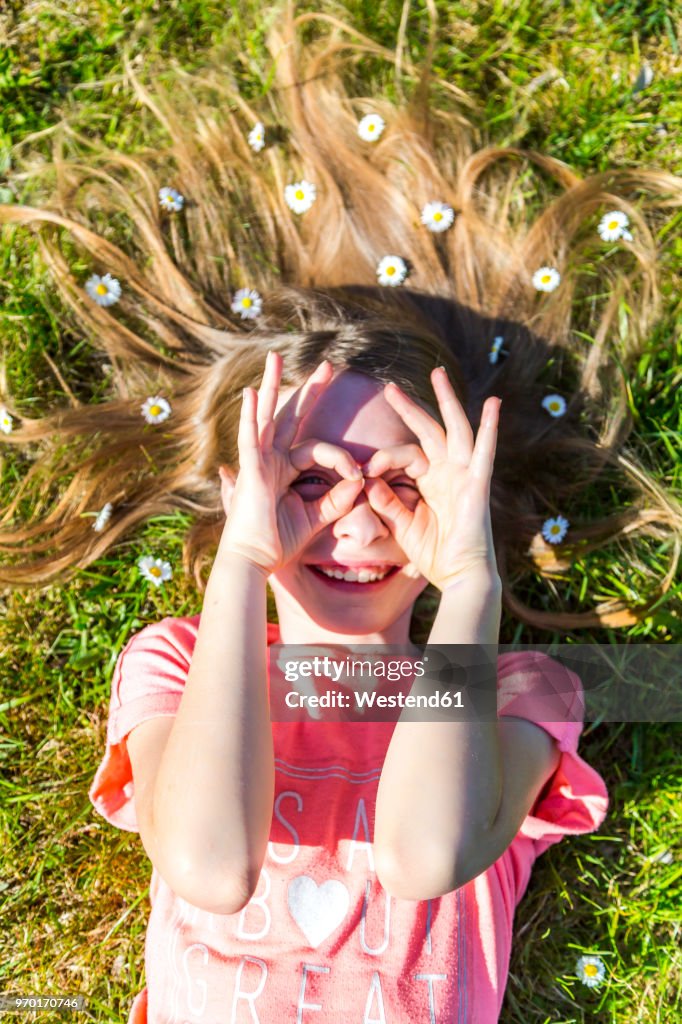 Smiling girl lying on grass in spring with daisies on hair forming spectacles with fingers