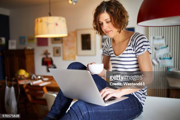 portrait of mature woman sitting on kitchen table using laptop - 40s laptop stockfoto's en -beelden