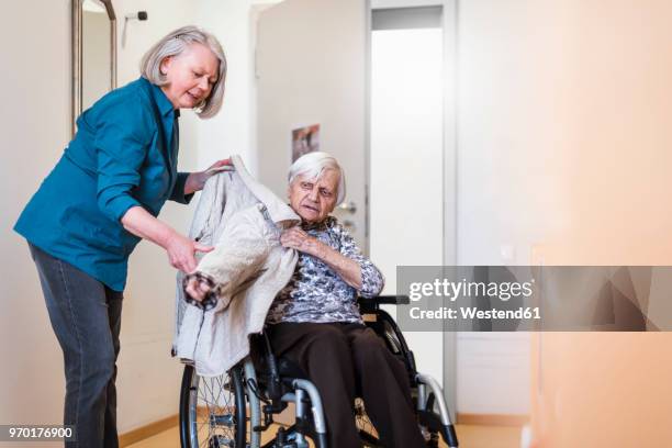 woman taking care of old woman in wheelchair putting her jacket on - fürsorge stock-fotos und bilder