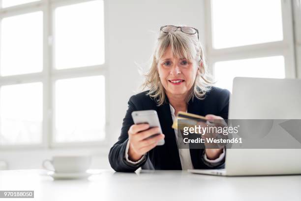 mature businesswoman with smartphone, credit card and laptop at desk in the office - webshopper stock-fotos und bilder