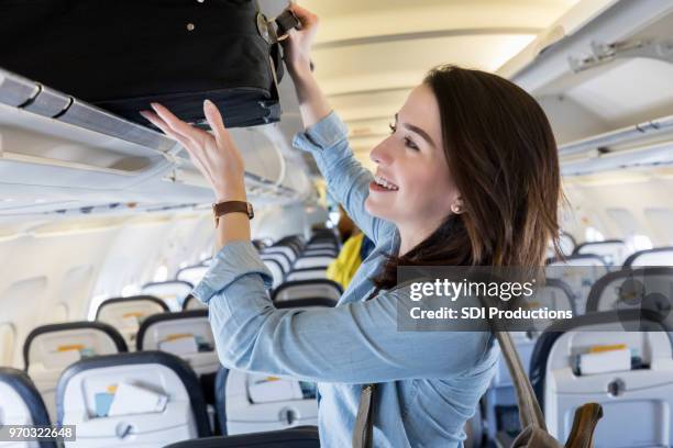 young woman puts carry on bag in overhead bin of airliner - heavy backpack stock pictures, royalty-free photos & images