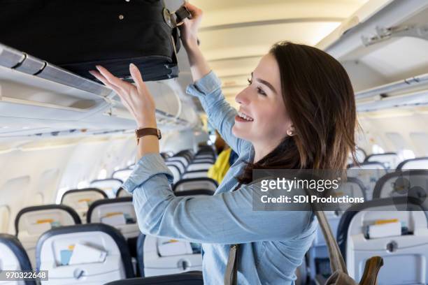 young woman puts carry on bag in overhead bin of airliner - carry on bag imagens e fotografias de stock