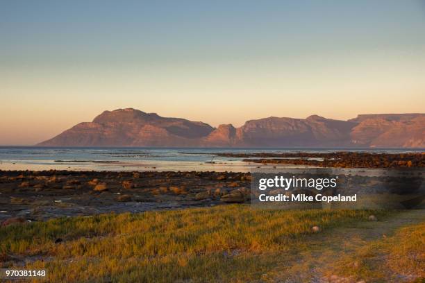 a sunset view across the rocks and kelp beds of kommetjie towards the karbonkelberg and sentinel mountains above hout bay on the shores of the cold atlantic ocean in the western cape of south africa. - south atlantic ocean stock pictures, royalty-free photos & images