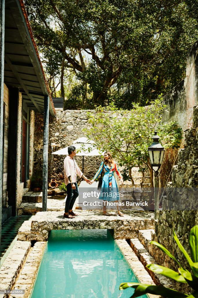 Smiling couple holding hands and walking across bridge over pool in courtyard of boutique hotel