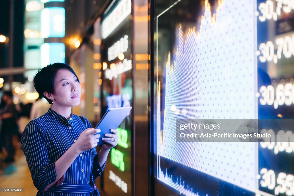 Asian businesswomen checking stock market data on tablet before Hong Kong financial display board