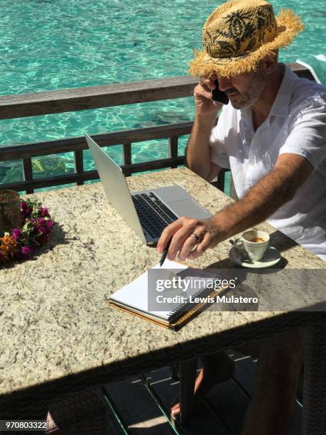 business retreat man sat outside on a wooden deck of a tropical overwater bungalow working remotely and wirelessly on computer whilst taking hand written notes from smartphone conversation - sattel bildbanksfoton och bilder