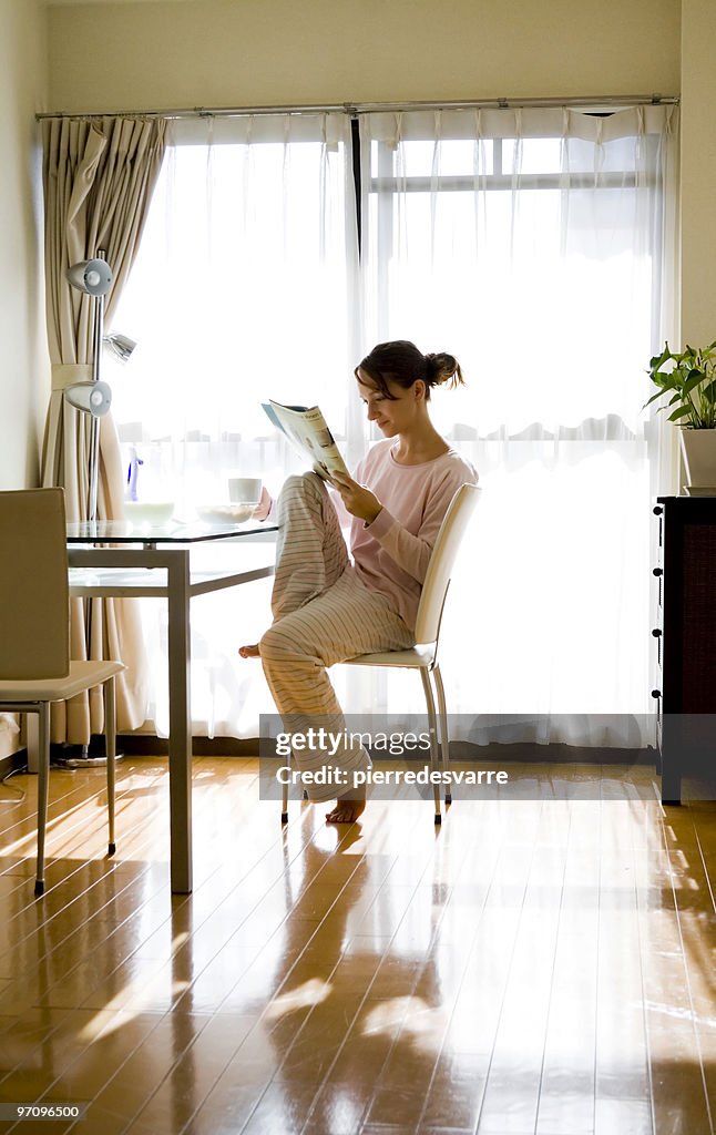 Woman sitting and relaxing by sunny window with magazine