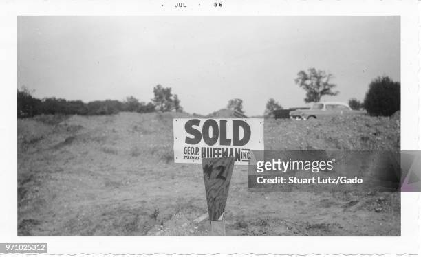 Black and white photograph, showing a sign with the words "Sold, GEO P Realtors, Huffman Inc" at a construction site with mounds of dirt in the...