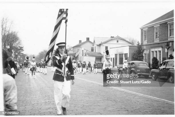 Black and white photograph, showing a parade, led by a uniformed boy holding an American flag, with majorettes and a marching band in the background,...