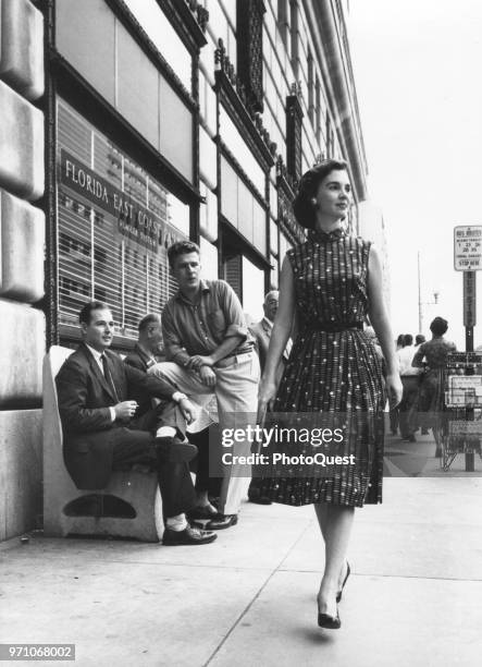 Pair of men on a bench stare as a woman walks past them, Florida, 1959. The building at left houses an office of the Florida East Coast Railway,...