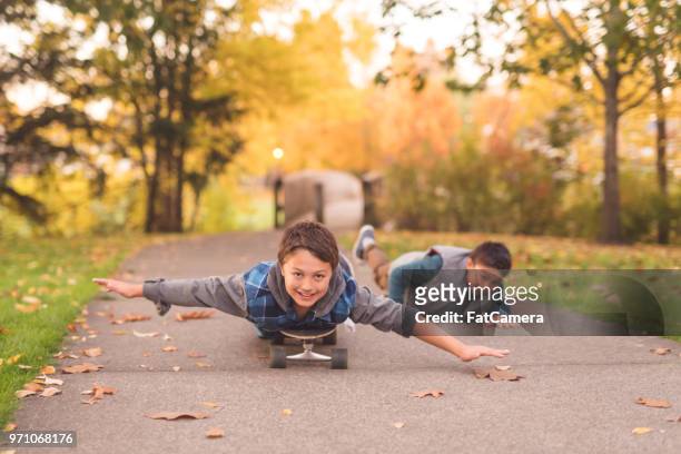 two elementary-age brothers ride skateboards through a park - november 2 2017 stock pictures, royalty-free photos & images