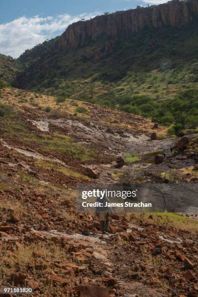 senior hiker walks down winding track through the rocks towards her companions high up in the hills, etendeka, namibia (model release) - james strachan stock pictures, royalty-free photos & images