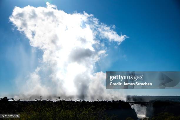 spray rises high above the victoria falls against an azure blue sky since the waters of the zambezi river are in full flood, with the famous victoria falls bridge bottom right, zimbabwe - james strachan stock pictures, royalty-free photos & images