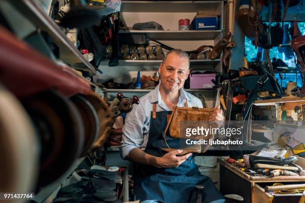 photo of mature cheerful shoemaker in workshop holding shoes and bag. looking at camera. - camera bag stock pictures, royalty-free photos & images