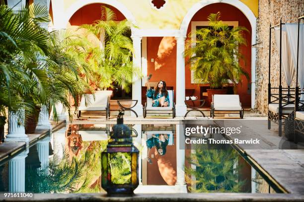 woman sitting in lounge chair by hotel pool taking selfie with smartphone - merida mexico stock-fotos und bilder