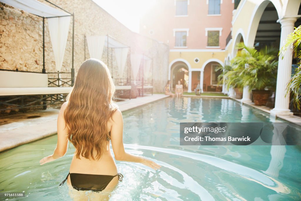 Woman wading into pool in courtyard of hotel while friends sit on edge