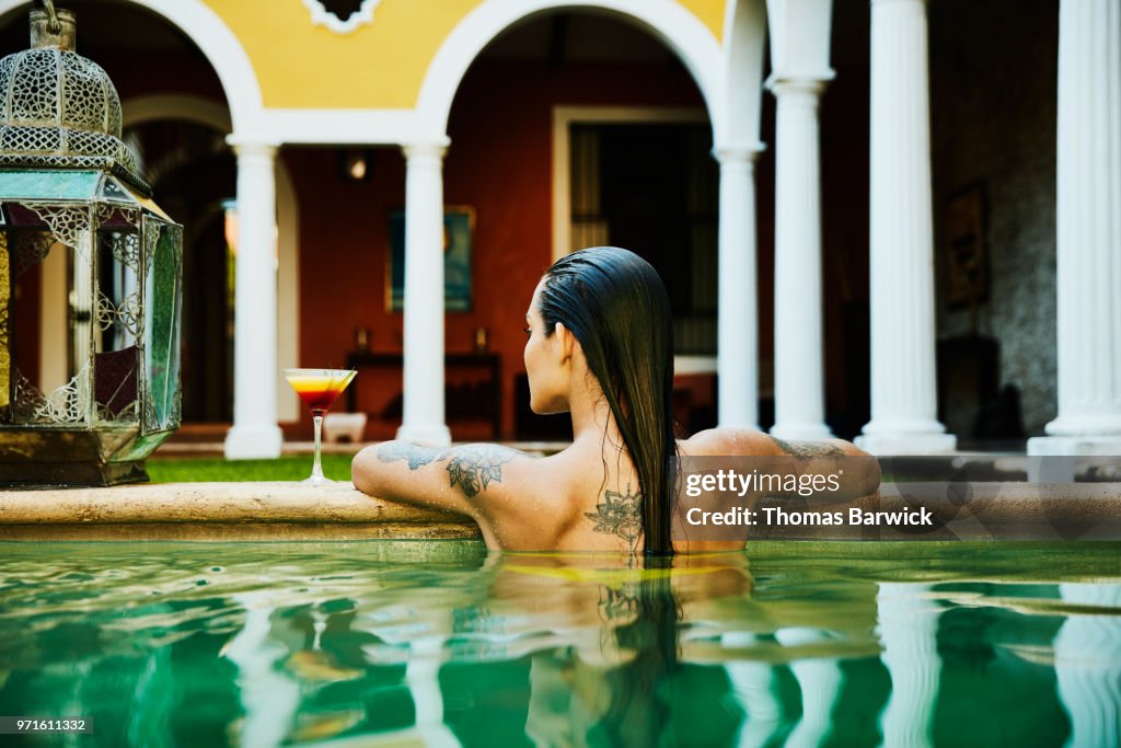 Rear view of woman relaxing on edge of pool with drink in courtyard of boutique hotel