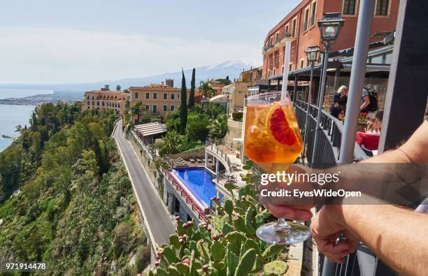 Man is holding an aperitif drink Aperol Sprizz with view to Mount Etna and the ocean on April 8, 2018 in Taormina, Sicily, Italy.