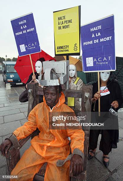Man sits in a fake electric chair as people wearing white masks hold banners reading "Halt to death penalty", during a protest to denounce the death...
