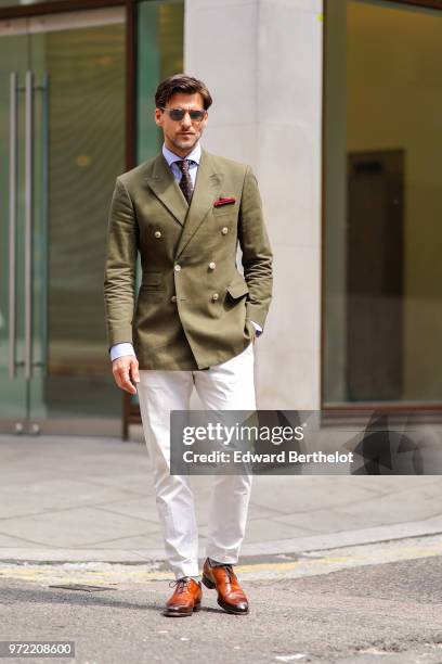 Johannes Huebl wears a green khaki blazer jacket, sunglasses, white pants, brown leather shoes, a blue shirt, a tie, during London Fashion Week Men's...
