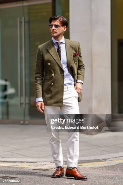 Johannes Huebl wears a green khaki blazer jacket, sunglasses, white pants, brown leather shoes, a blue shirt, a tie, during London Fashion Week Men's...