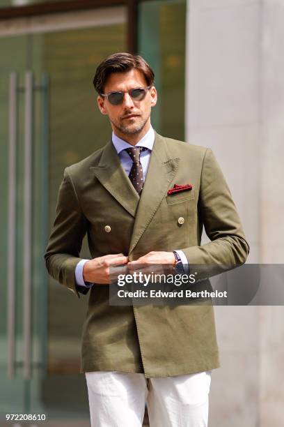 Johannes Huebl wears a green khaki blazer jacket, sunglasses, white pants, brown leather shoes, a blue shirt, a tie, during London Fashion Week Men's...