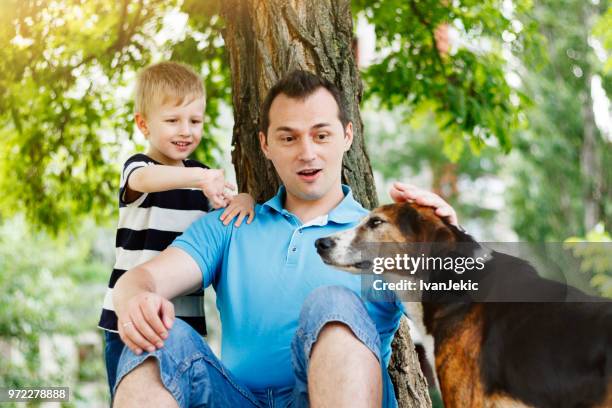 father and son cuddling dog in nature - ivanjekic stock pictures, royalty-free photos & images