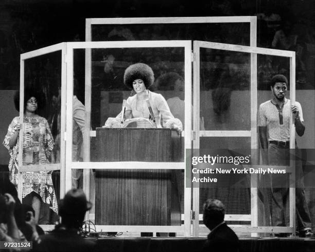 Angela Davis speaking at Madison Square Garden behind a four-sided, 7-foot high bulletproof glass shield.