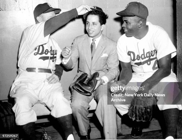 King Faisal II of Iraq, in the Brooklyn Dodgers' dugout, is crowned with a baseball cap by Chuck Dressen while Jackie Robinson holds an autographed...