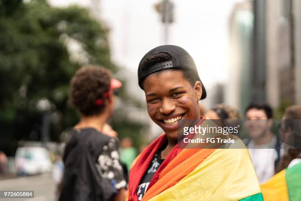 portret van een jonge man met regenboogvlag met vrienden op de achtergrond bij gay parade - lgbtqi stockfoto's en -beelden