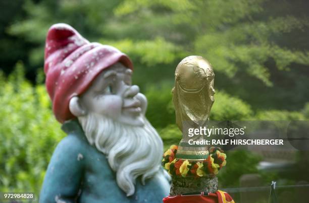 Mockup of the FIFA Football World Cup trophy decorated with a flower chain in the colors of Germany stands next to a garden gnome in a garden in...