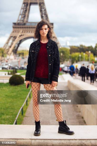 Model Mckenna Hellam poses in front of the Eiffel tower and wears an embellished denim jacket, red sweater, black shorts, fishnet tights and black...