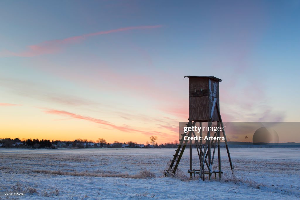 Raised hide for hunting roe deer at sunrise in snow covered field in winter.