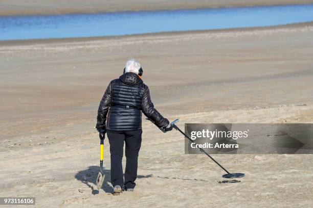 Elderly woman with metal detector beachcombing on sandy beach along the North Sea coast in winter.