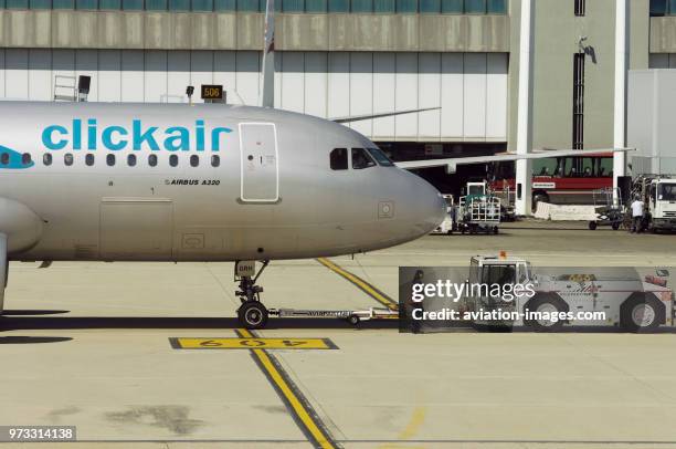 Forward fuselage of a clickair Airbus A320-200, a tug with a towbar, an airport worker wearing an orange tabard and ear-defenders with a jetway and...