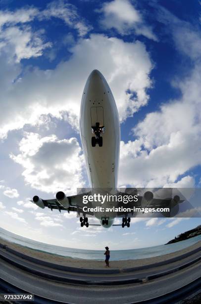 A man in a hat cowering as an A340 passes overhead on low final-approach landing over the sea at Maho Beach with clouds.