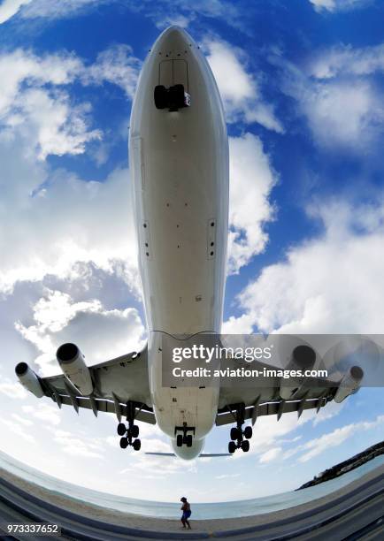 A man in a hat cowering as an A340 passes overhead on low final-approach landing over the sea at Maho Beach with clouds.