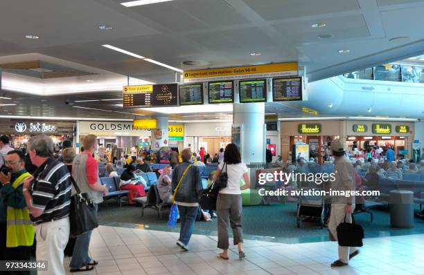 Passengers with baggage looking at the flight departures screen with Fat Face, Goldsmith's, Clarol Studio and Harrods of Knightsbridge Duty Free...