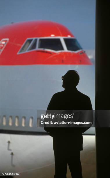 Man wearing spectacles standing by a window in the departure-lounge with the nose of a Northwest Airlines Boeing 747 parked outside.