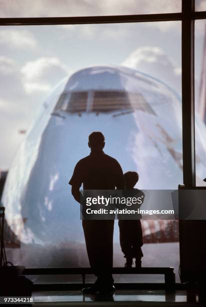 Man and child standing at the window in the departure-lounge with the nose of a Boeing 747 parked outside.